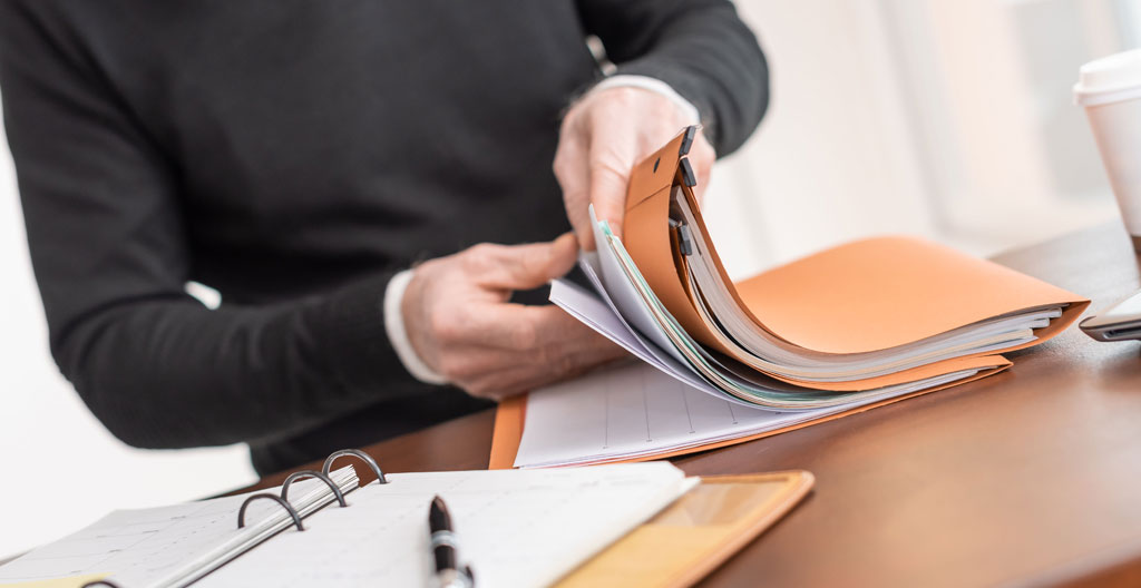 Une personne vue de dos assise à un bureau simple en train de remplir des formulaires administratifs français, plusieurs documents CERFA étalés, stylo en main, dans un bureau à domicile contemporain éclairé par une fenêtre